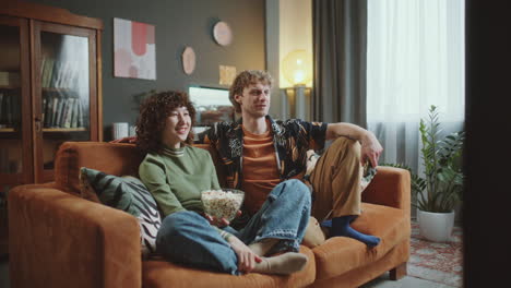young couple watching movie on tv, sharing popcorn and laughing at home