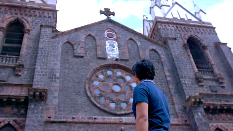 Young-man-looks-at-old-stone-church-in-Ecuador,-slow-pan-from-behind