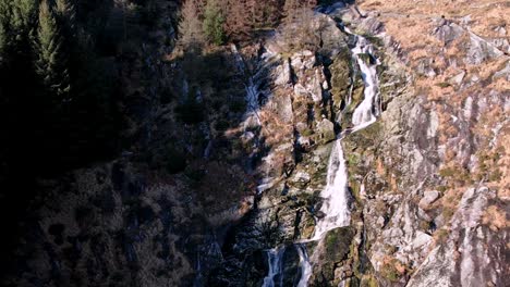 Una-Vista-Aérea-De-Retroceso-De-La-Cascada-Glenmacnass-Que-Cae-80-Metros-Por-El-Valle-Del-Río-Glenmacnass-En-Las-Montañas-Wicklow-Cerca-De-Laragh-Que-Desemboca-En-El-Mar-En-Arklow-Cerca-De-La-Carretera-Tr115