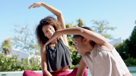 happy biracial mother and daughter practicing yoga stretching in sunny garden, slow motion