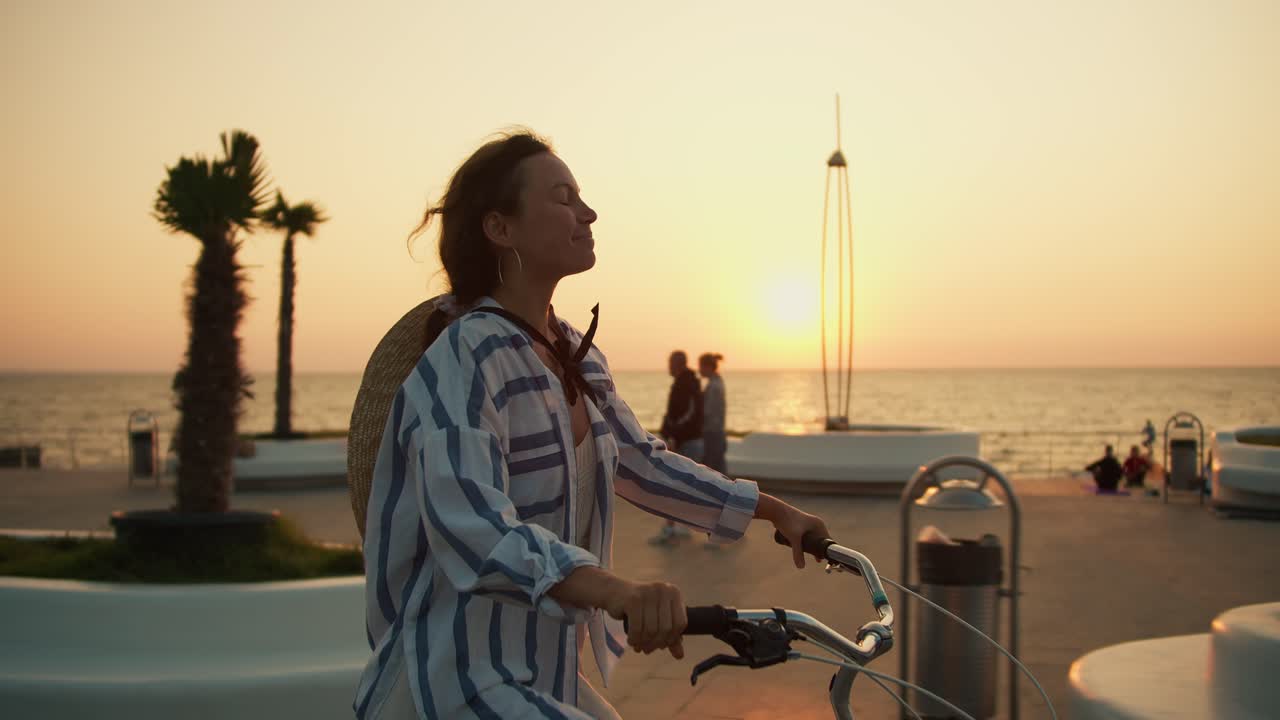 Happy brunette girl in a striped shirt rides a bicycle along the sea on the beach in the morning at dawn. Morning bike ride on the beach at sunrise
