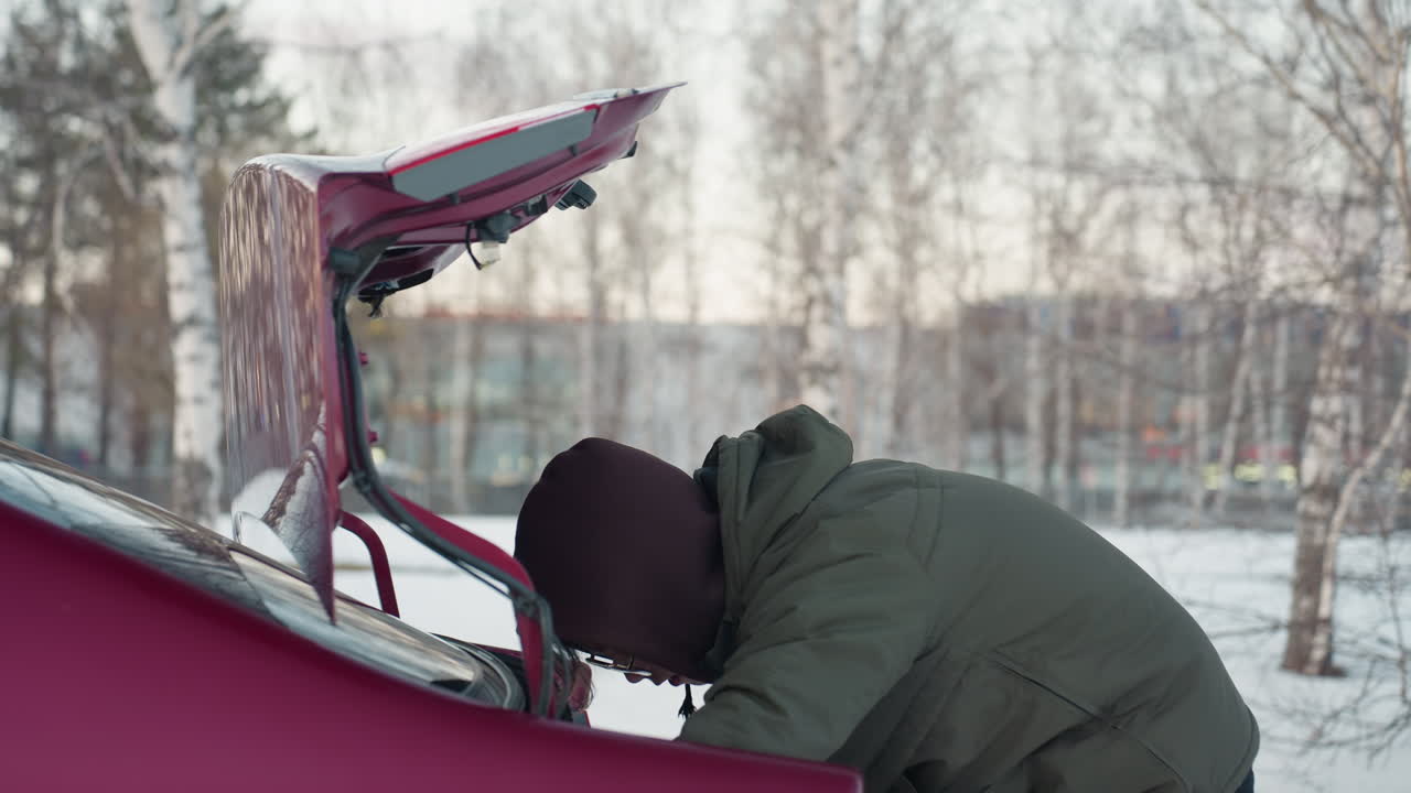 Young man busy with something in car boot, surrounded by nature, with trees in the background, outdoor scene capturing action and focus on vehicle in peaceful natural environment