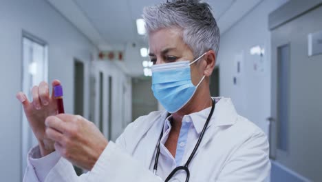 Portrait-of-caucasian-female-senior-doctor-wearing-face-mask-holding-blood-sample-in-hospital