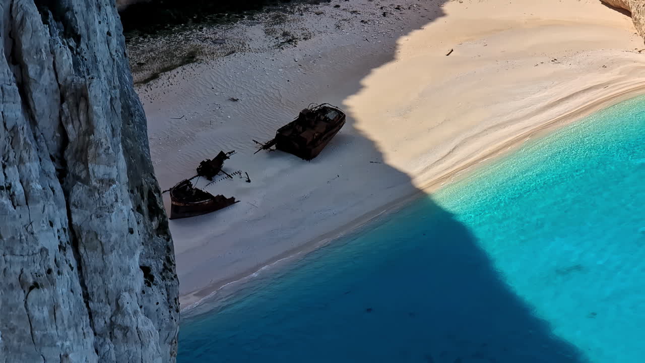 Skibsvrag i skyggen af en stejl klippe sunket under gyldent hvidt sand på strand med klart tropisk blåt vand ved Navagio.