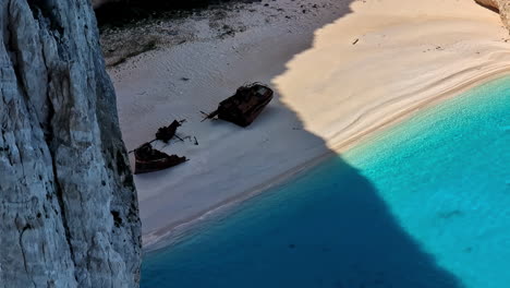 Shipwreck-in-shade-of-steep-clif-sunk-below-golden-white-sand-on-beach-with-clear-tropical-blue-waters-of-Navagio