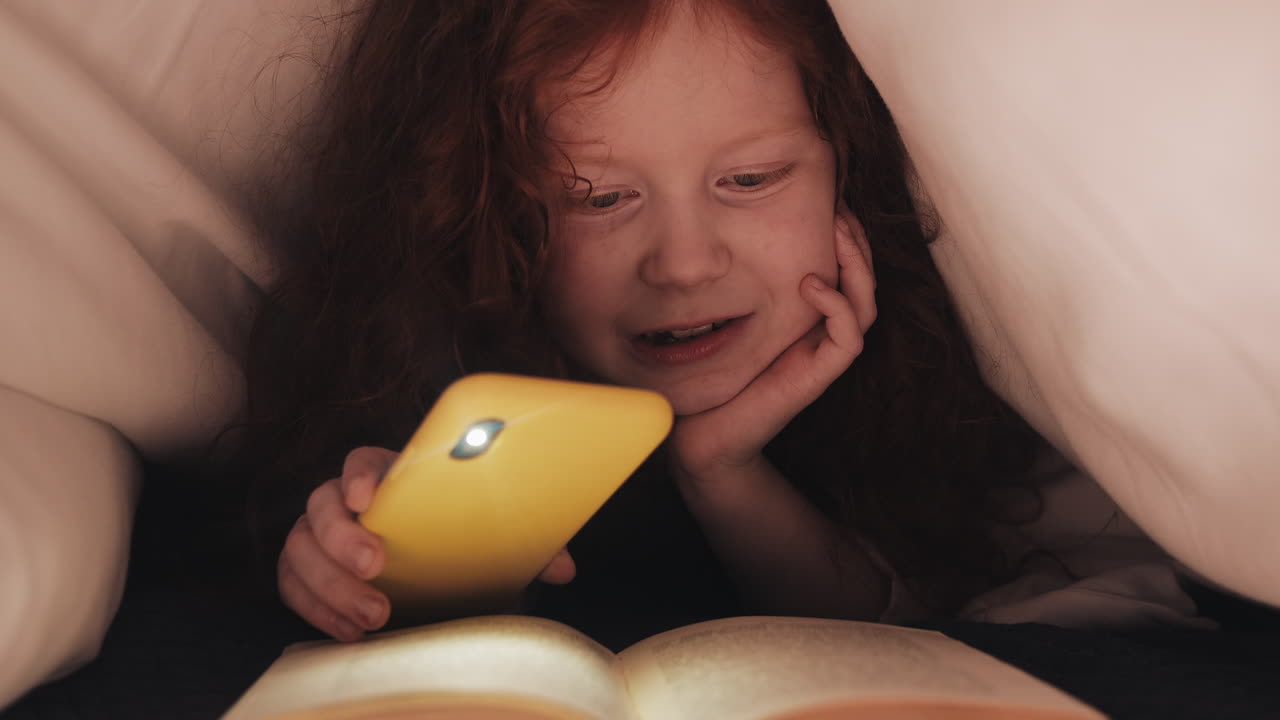 Redhead girl reading on her phone under the covers at night