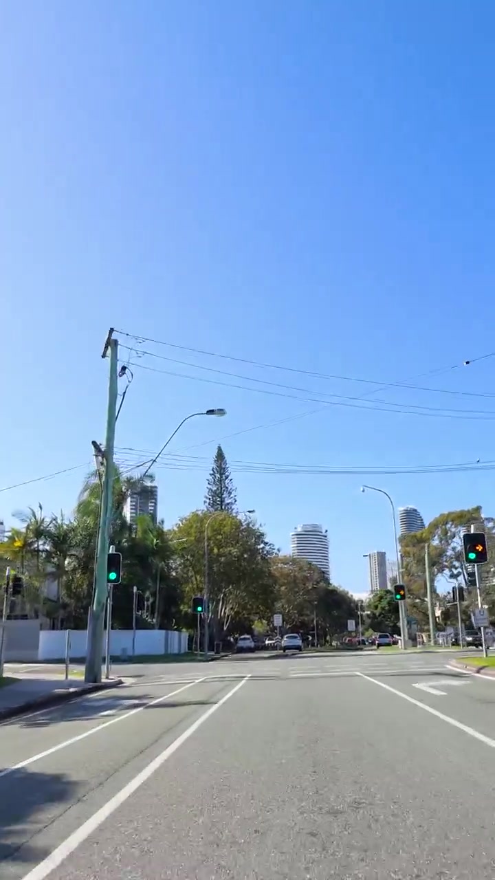 Vue depuis un véhicule en mouvement sur une route asphaltée sous un ciel bleu, avec des arbres et des gratte-ciel en milieu suburbain.