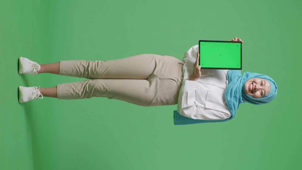 cuerpo lleno de mujer musulmana asiática sonriendo y mostrando la tableta de pantalla verde a la cámara mientras está de pie en el estudio de fondo de pantalla verde