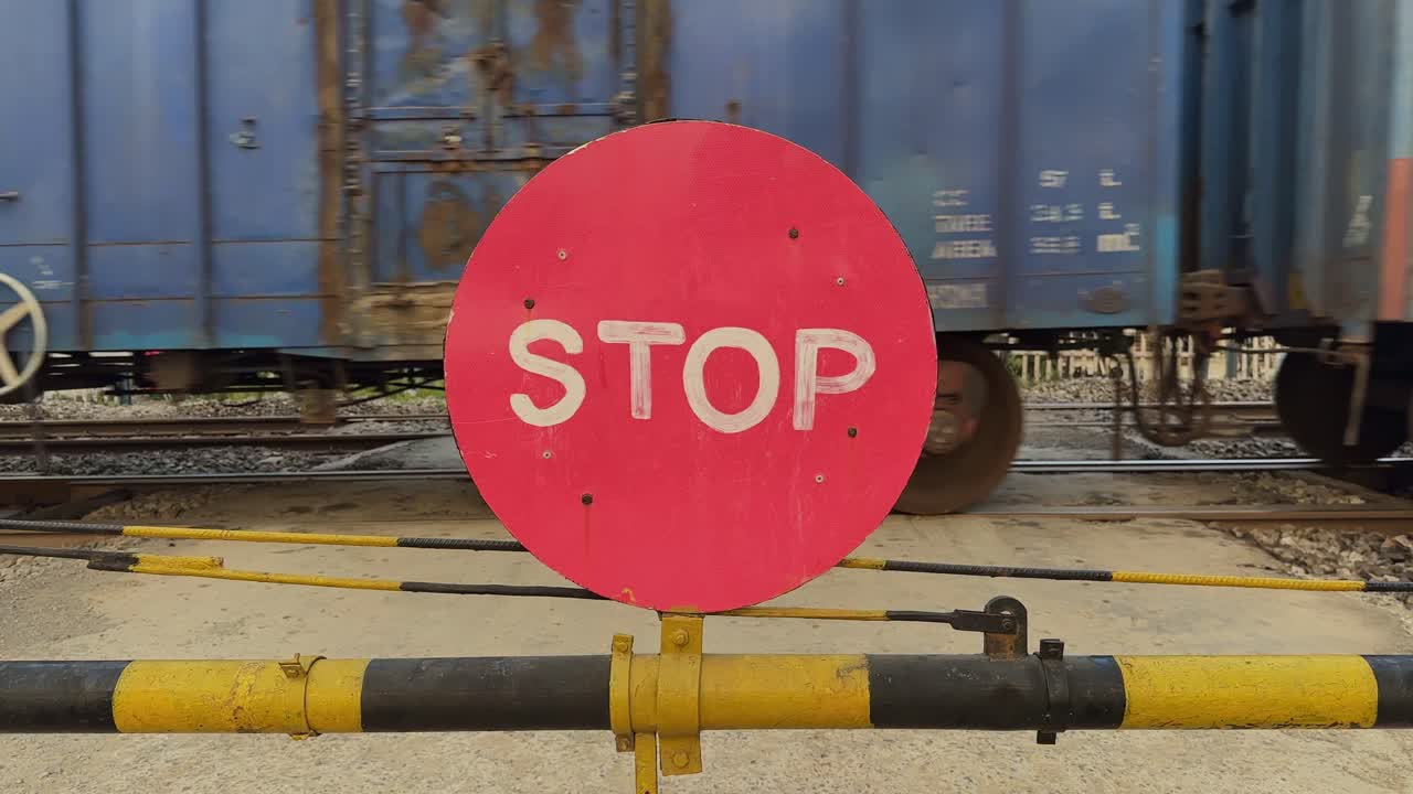 Close-up view of a red STOP sign fixed to the lowered yellow barrier as a blue freight train rushes past in the background with strong motion blur