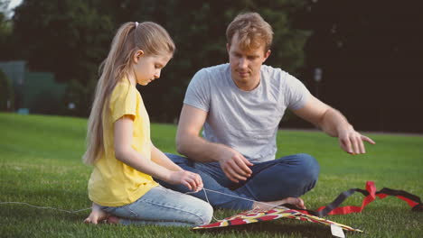 Padre-E-Hija-Arreglando-Cometas-Sentados-En-Una-Hierba-Verde-En-El-Parque