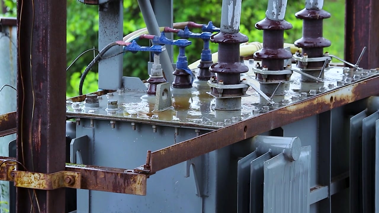 Sottostazione di trasformazione elettrica che mostra isolatori bagnati e telaio metallico arrugginito con acqua piovana che gocciola dalle alette di raffreddamento, apparecchiature elettriche industriali