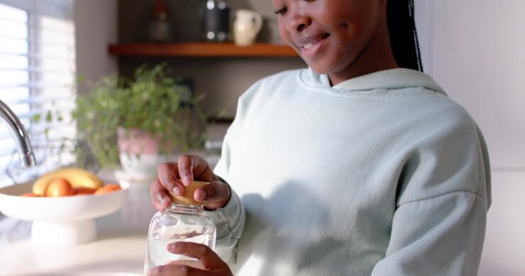 African teenage woman tapping wooden cap, lifting twisting sealing water bottle at counter smiling