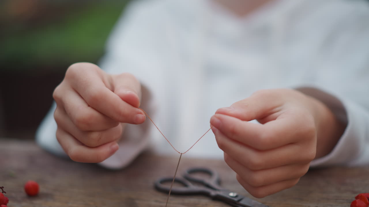 Closeup small hands threading fine thread through bead, scissors on wooden table, outdoor garden blur, white child concentrating, patient jewelry making, gentle natural light, careful fingerwork, slow