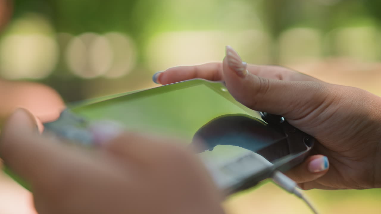 Mujer negra jugando a la consola portátil en el parque, jugadora relajada disfrutando de la luz del sol veraniega, movimiento del pulgar y detalles de las uñas, reflejo del dispositivo portátil, bokeh suave de la naturaleza, hobby de ocio, poca profundidad cinematográfica