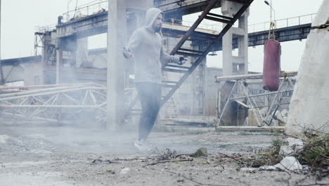 close-up view of caucasian man with beard in a grey hoodie jumping rope outdoors an abandoned factory on a cloudy morning