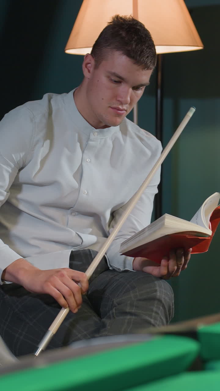 jugador de billar con camisa blanca y pantalones grises se sienta en una silla blanca, leyendo un libro mientras sostiene el palo de la clave con expresión enfocada. la iluminación cálida mejora la atmósfera reflexiva e intelectual