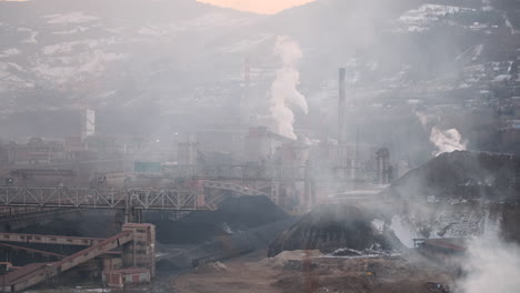 smoke billows from towering stacks at an industrial complex nestled in a snowy mountainous region