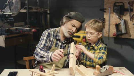 caucasian senior man carpenter teaching his small cute grandson how to work with wood in workshop at desk