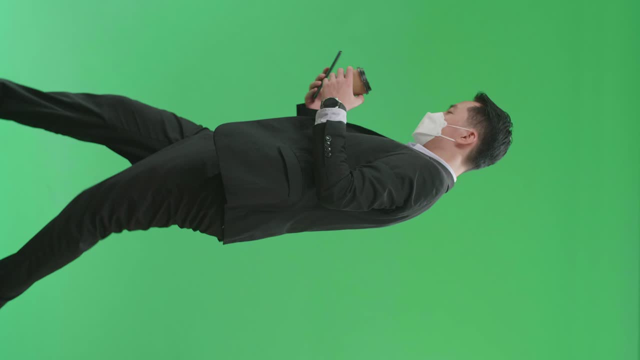 Side View Of Asian Business Man Use Mobile Phone And Drink Coffee While Walking On Green Screen Chroma Key, Wear Medical Mask