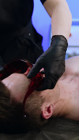 caucasian man lies on the couch in beauty salon. cosmetologist comes up and puts protective goggles on client. vertical video