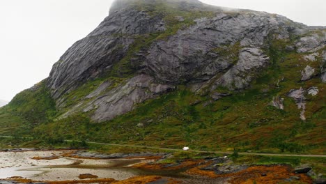 Aerial-view-of-a-white-camper-van-on-a-coastline-road-at-the-foot-of-a-majestic-mountain,-Lofoten-Islands,-Norway