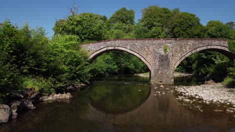 Under-an-old-stone-arch-bridge,-trees-and-small-river-captured-by-drone