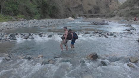 three men hikers trekking across river creek water flowing rocks boulder low flying aerial orbiting right around people as they cross river