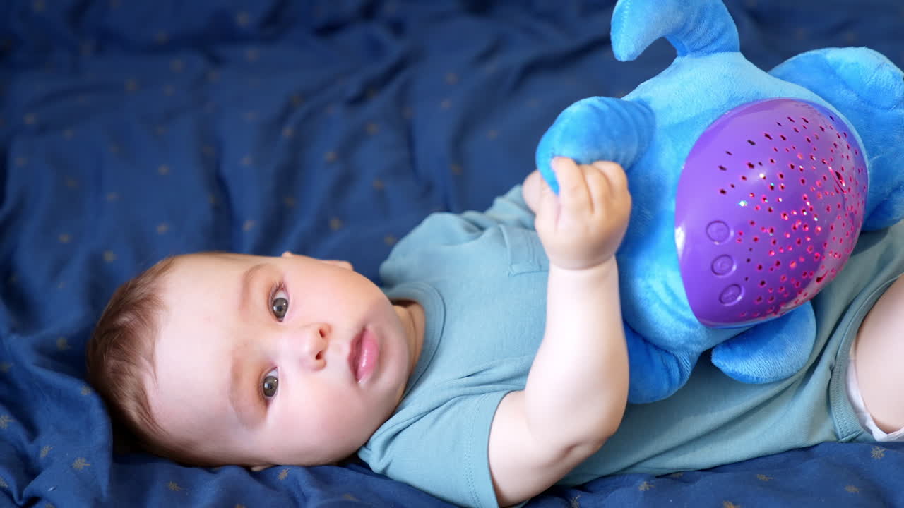 Beautiful Caucasian baby boy with a soft toy in hands. Nice kid distracts from a toy looking at camera and then turns to his toy again. Close up. Blue backdrop.