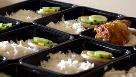 a row of nasi lemak in lunch boxes container while hands put a hot crispy chicken roasted on it