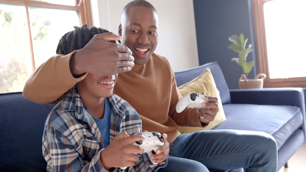 Happy african american father and son playing video games and fooling around at home, slow motion