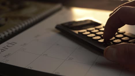 Hands-using-calculator-on-March-calendar-panning-shot-warm-background