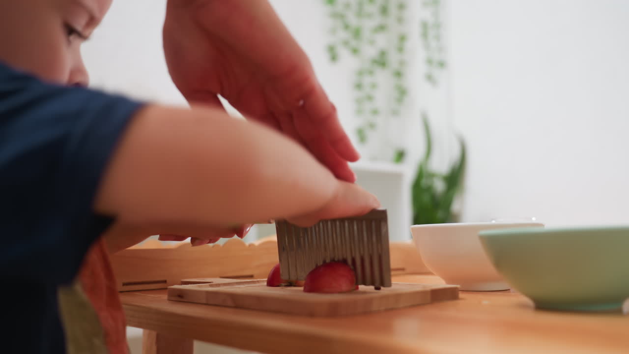Little boy uses chopper to cut apple on wooden board while instructor reaches to guide, focusing on safe cooking practice and learning food preparation in bright kitchen setting