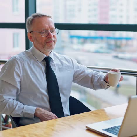 senior freelancer using laptop working from home. smiling grey-haired man in glasses typing on computer and drinks coffee enjoying distant job.