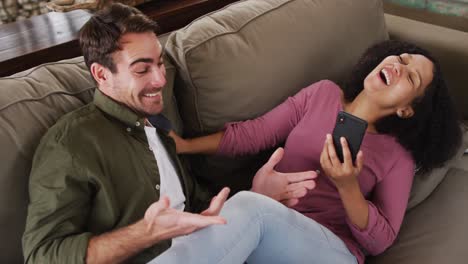pareja de raza mixta sonriendo mientras miran en el teléfono inteligente juntos en el sofá en la casa de vacaciones