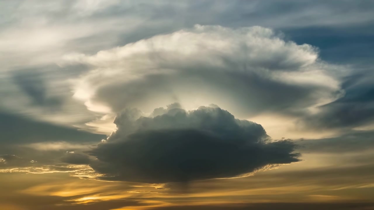 Massive cumulonimbus cloud fills wide sky with dense dark base, towering cauliflower top, bright backlit anvil glow, soft rim light, layered blue atmosphere, and warm horizon light