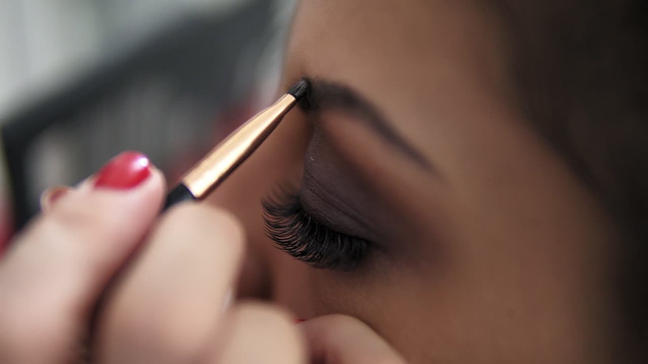 Close Up view of the makeup artist's hands using brush to paint eyebrows for a model with false lashes. Slow Motion shot