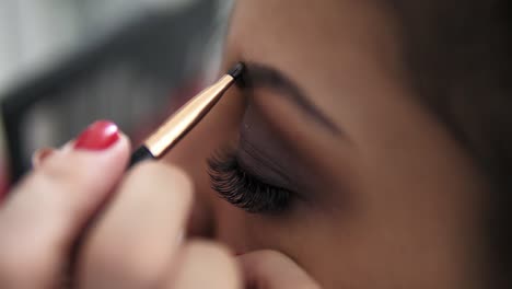 close up view of the makeup artist's hands using brush to paint eyebrows for a model with false lashes. slow motion shot