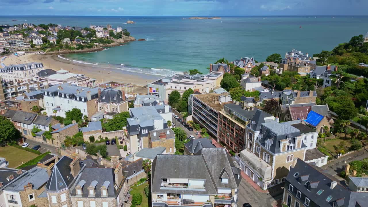 Dinard coastline with historic seaside architecture, sandy beaches and calm turquoise waters of the English Channel beneath a clear blue summer sky, aerial panoramic view, drone pulling away slow
