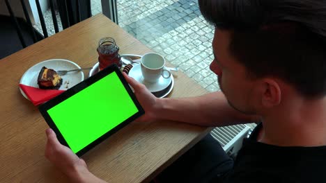 un hombre se sienta en una mesa con comida en un café y mira una tableta con una pantalla verde - primer plano desde arriba