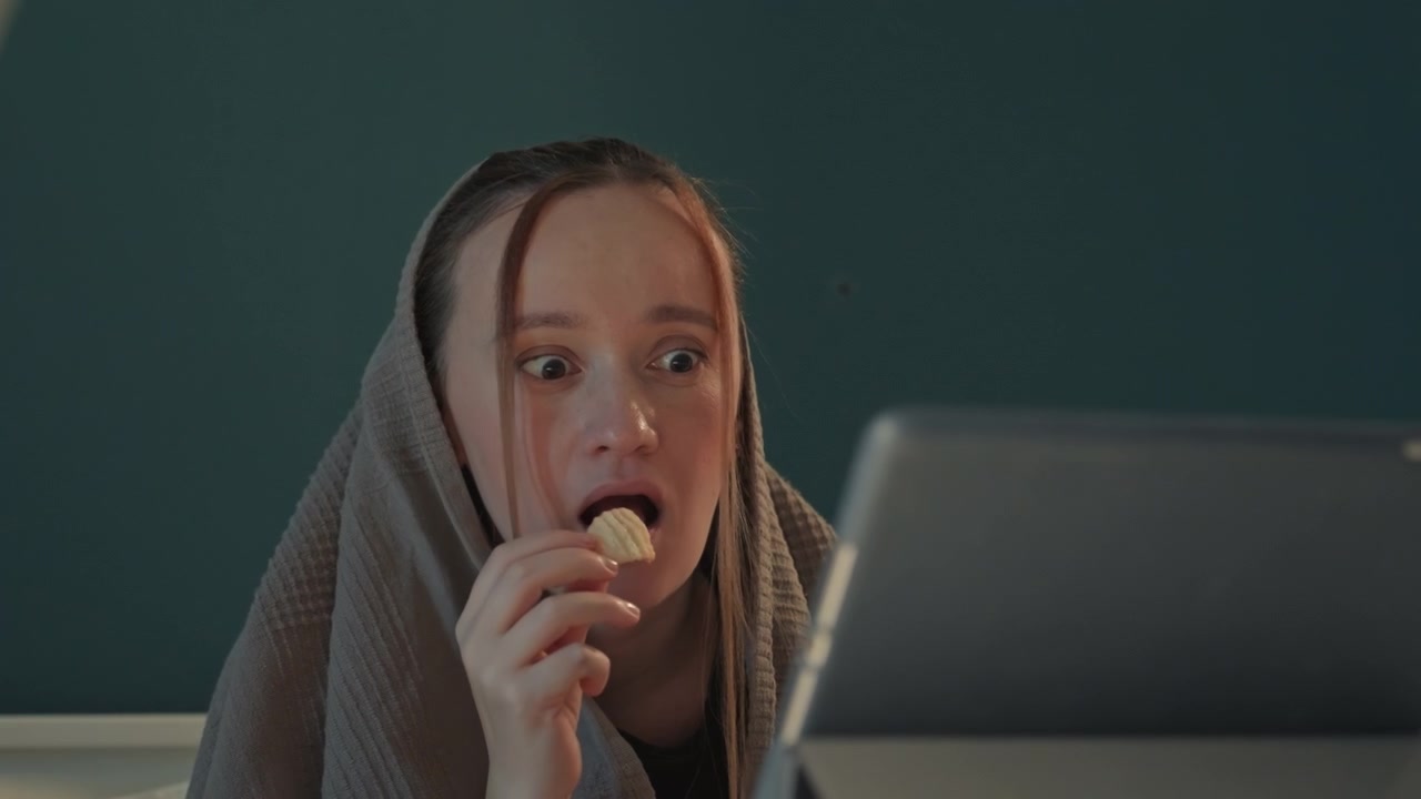 Ung kvinde med langt hår sidder ved et skrivebord, svøbt i et gråt tæppe. Kvinden spiser kartoffelchips, mens hun kigger mod kameraet. Bærbar computer og en blågrøn væg er synlige bag hende gennem hele scenen.