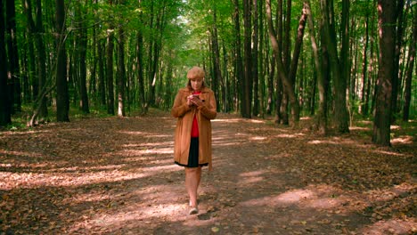 caucasian women with a jacket and hat looking into mobile and walking in the forest trail