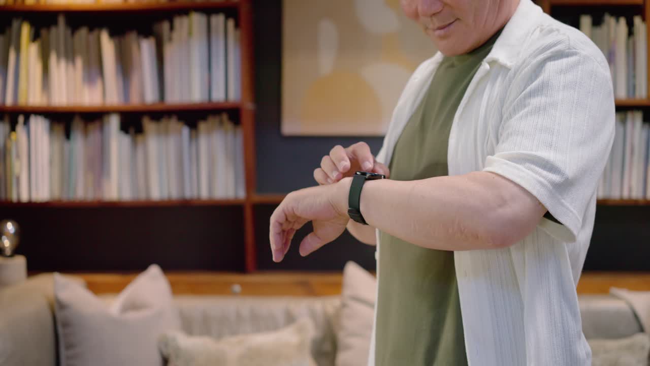 Senior Asian man raising wrist, checking, tapping smartwatch after alert in living room, copy space