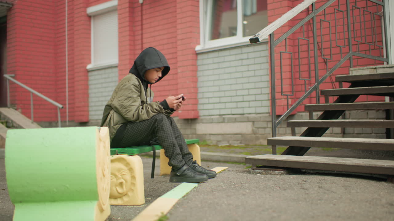 Side view of kid in hooded jacket seated on outdoor bench placing leg on wedge, focused on handheld device, with red brick building, stair railing, and muted urban environment in background