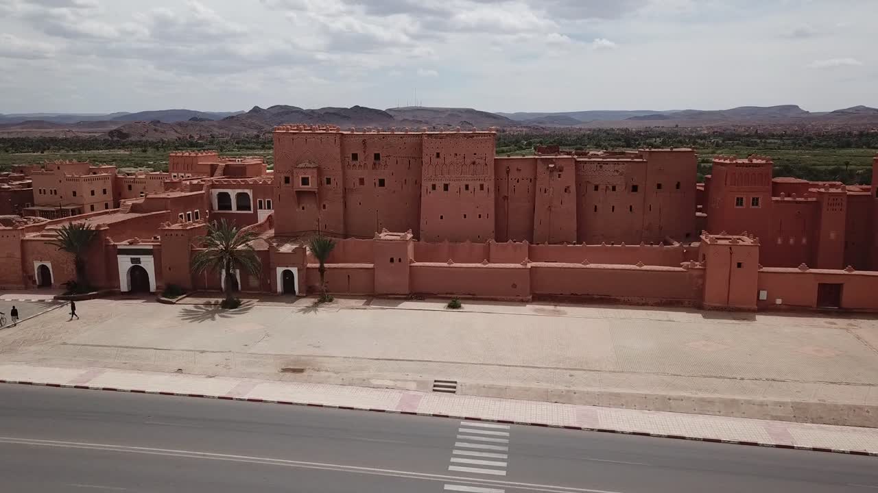 Vista frontale di una grande fortezza kasbah marocchina vicino a una strada asfaltata, che mostra l'architettura tradizionale in adobe in un contesto di città desertica. Ideale per temi di cultura, viaggi e patrimonio urbano.