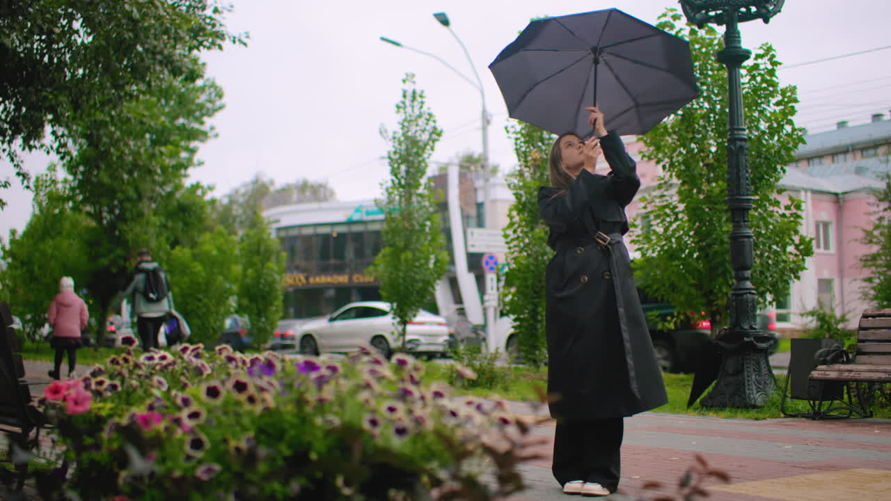 Young woman in long black coat struggles with umbrella in windy rainy weather on city street near benches and blooming flowers, surrounded by cars, buildings, and pedestrians creating urban lifestyle scene