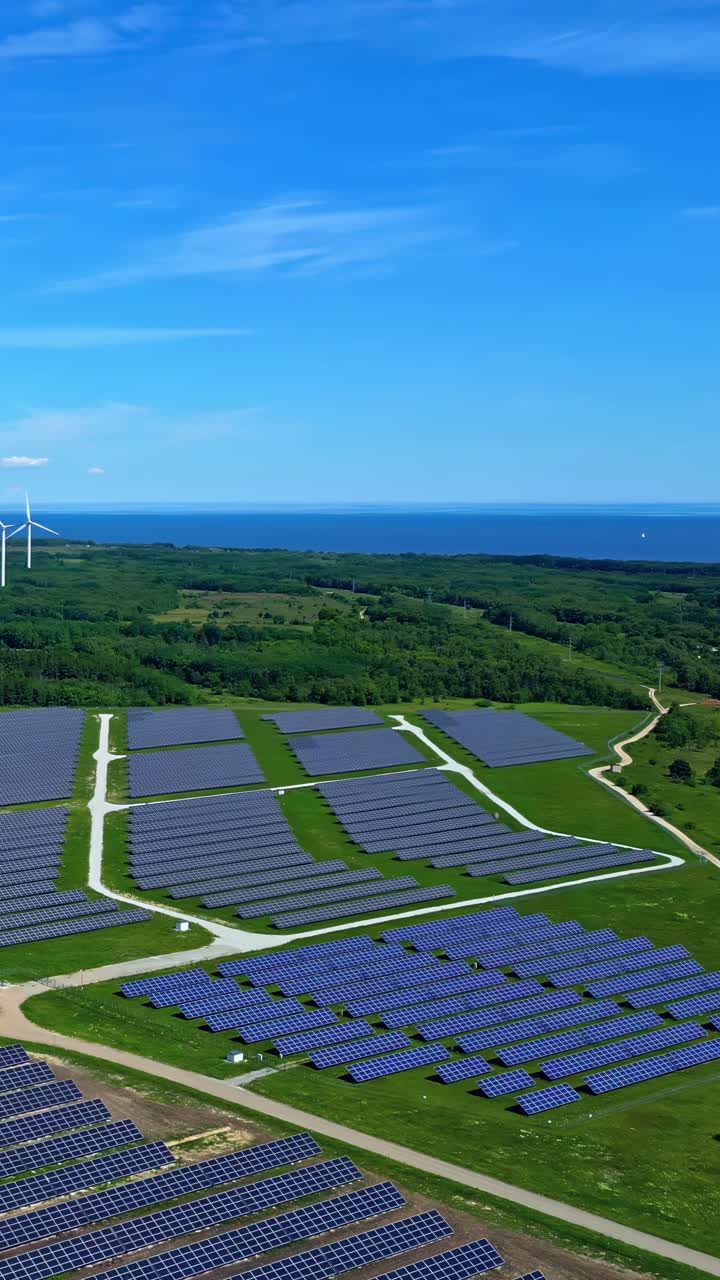 Wide view of solar park with forest, windmills, and ocean nearby in green energy landscape - vertical view