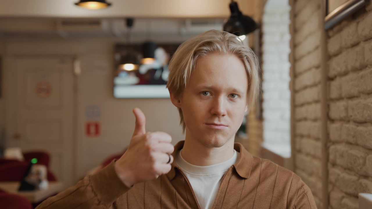 Nahaufnahme eines jungen Mannes mit kurzen blonden Haaren, der eine braune Jacke und ein weißes Hemd trägt, in einem Innenraum. Der Mann hält eine Tasse nahe an seinen Mund und macht dann eine Daumen-hoch-Geste, während er direkt blickt.