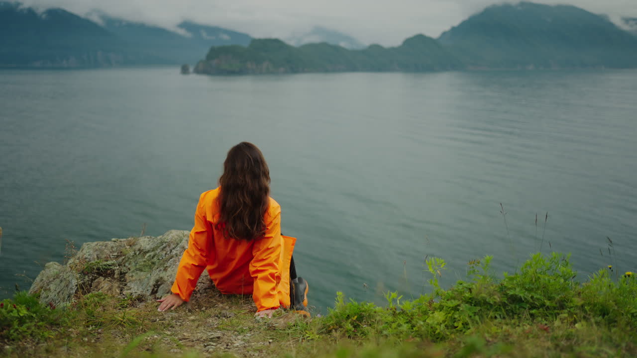 Woman in an orange raincoat sitting on a cliff overlooking the ocean and mountains