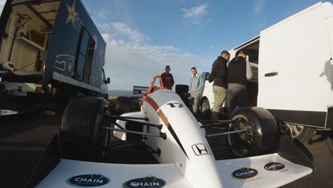 men walking closed to a white racing car parked between a van and a truck at the roadside on the hill in malta - gopro pan shot