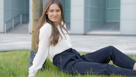 mujer de negocios relajándose, sonriendo contra un árbol fuera de su edificio de oficinas